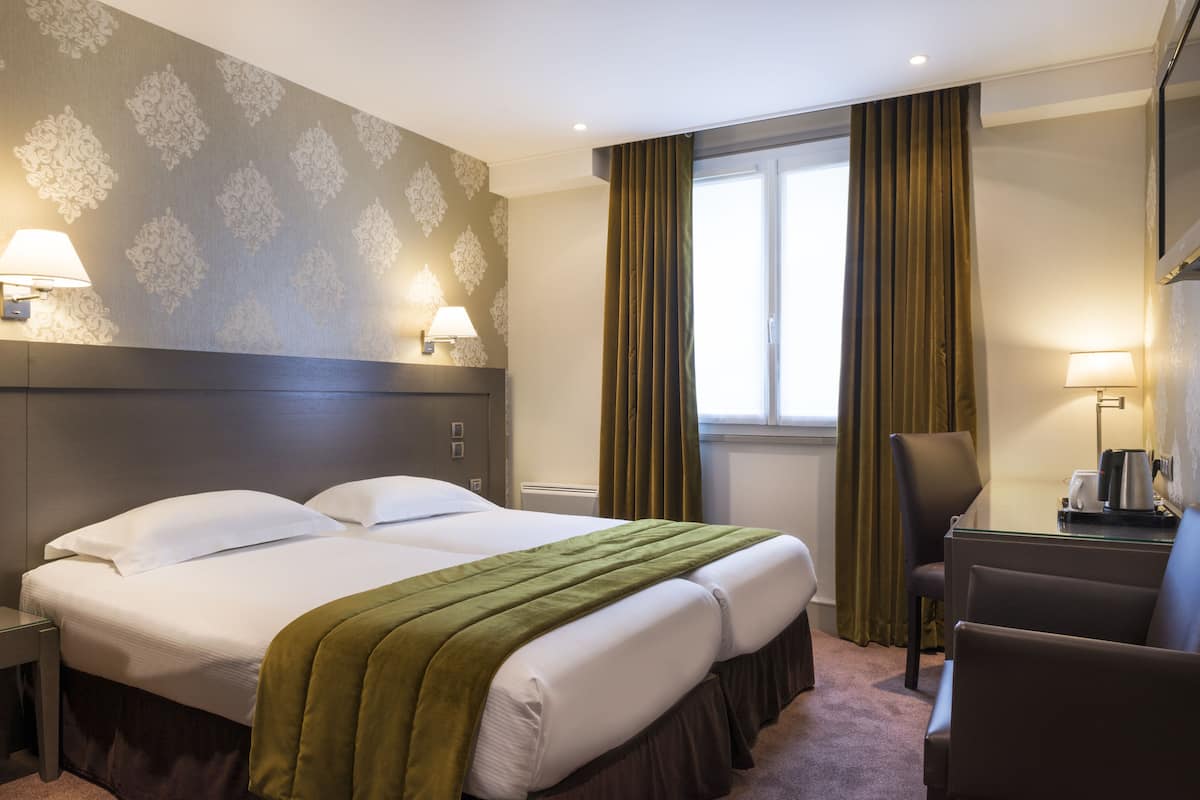 Habitación de hotel con dos camas, un escritorio con silla, una ventana con cortinas y lámparas fijadas a la pared.