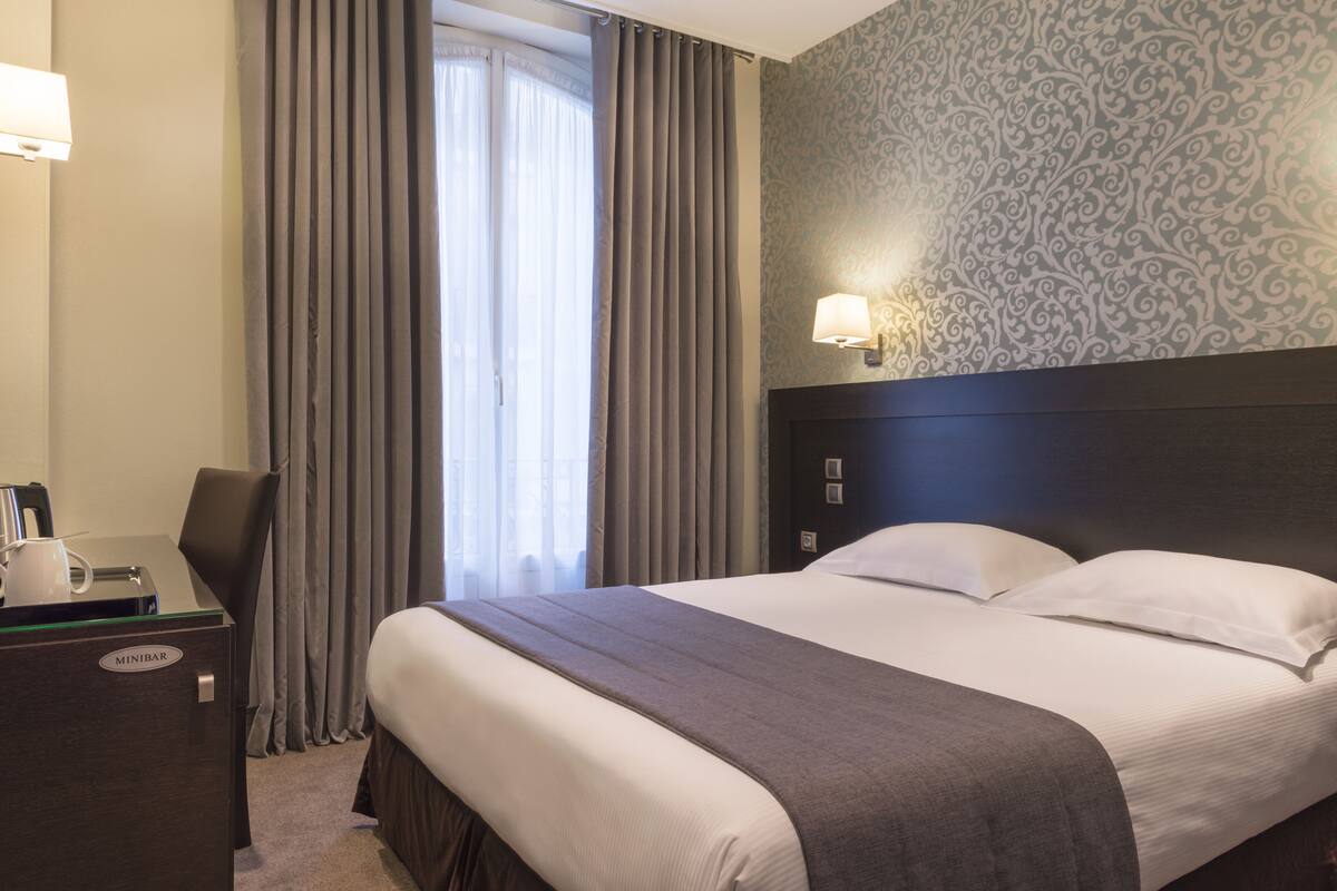 Una habitación de hotel con cama, escritorio, lámpara y ventana con cortinas.