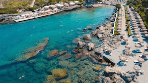 An aerial view of a coastal resort with clear turquoise waters, rocky shorelines, and a long promenade with sun loungers and umbrellas. - Atrium Platinum Luxury Resort Hotel & Spa (Rhodes)
