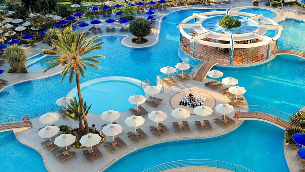 A large, multi-level swimming pool with sun loungers and umbrellas, surrounded by palm trees and a central bar area. - Atrium Platinum Luxury Resort Hotel & Spa (Rhodes)