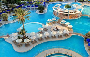 Una gran piscina de varios niveles con tumbonas y sombrillas, rodeada de palmeras y una zona de bar central.