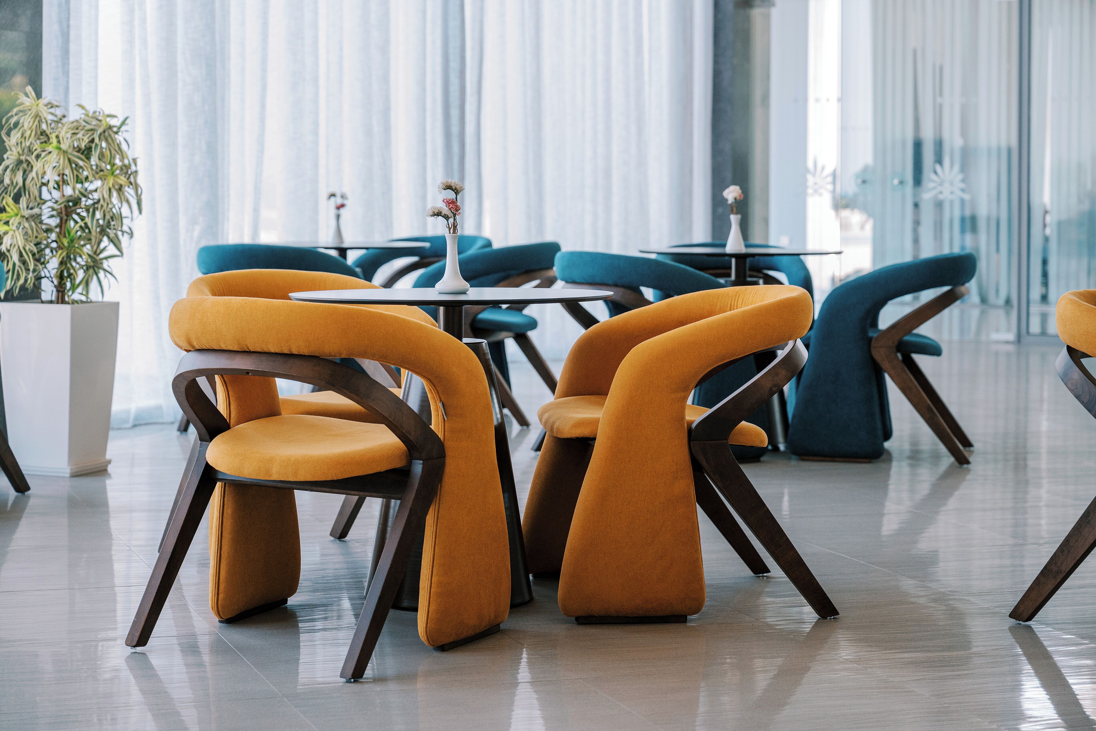 Un comedor moderno con sillas naranja y aguamarina, un jarrón blanco con flores y una planta en maceta.