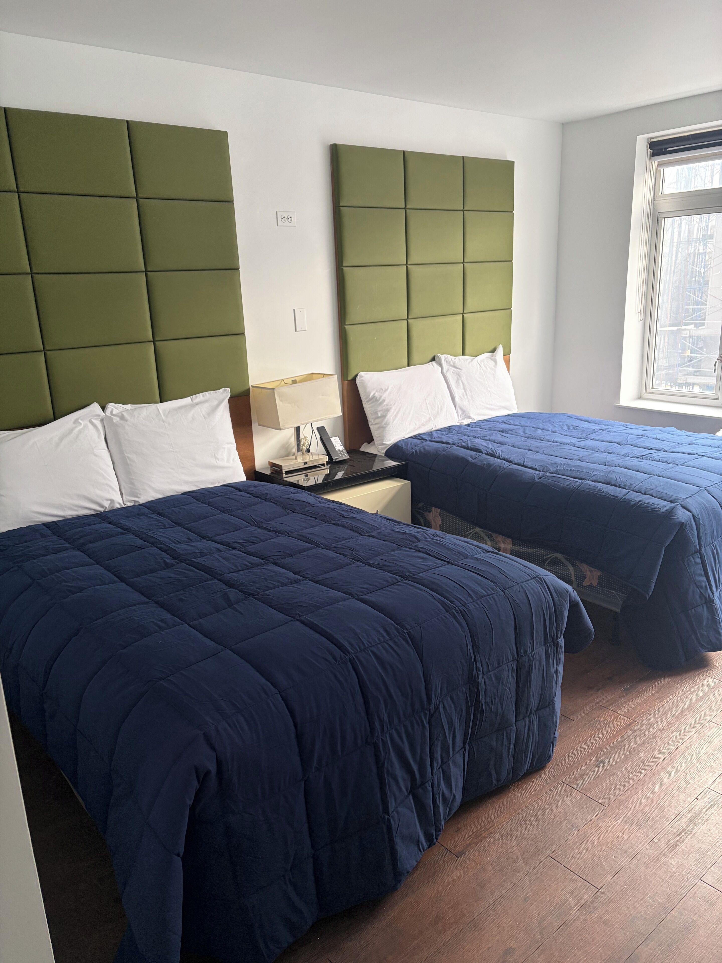 Een hotelkamer met twee bedden, elk voorzien van een groen hoofdeinde en witte beddengoed. Er is een raam met uitzicht op buiten, en een klein tafeltje met een lamp en telefoon.