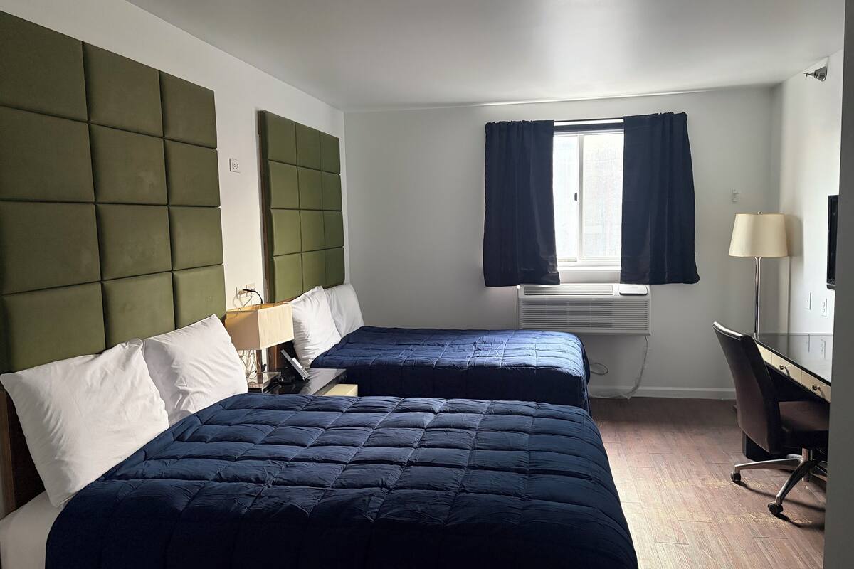 Een hotelkamer met twee bedden, een bureau, een stoel, een raam met gordijnen en een airconditioning.