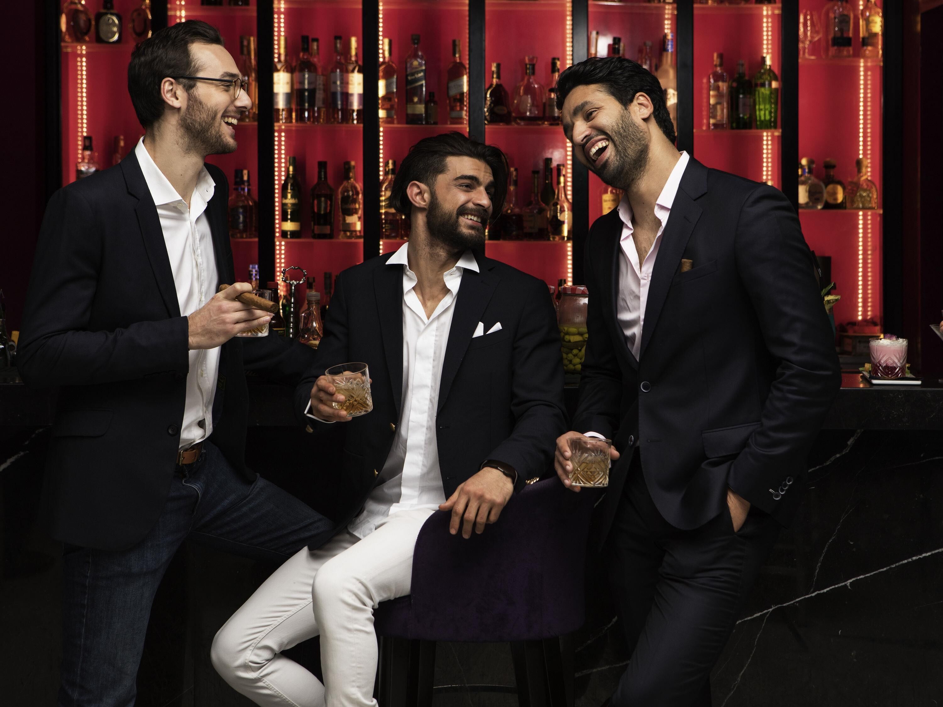 Tres hombres en trajes en un bar, con estanterías llenas de botellas al fondo.