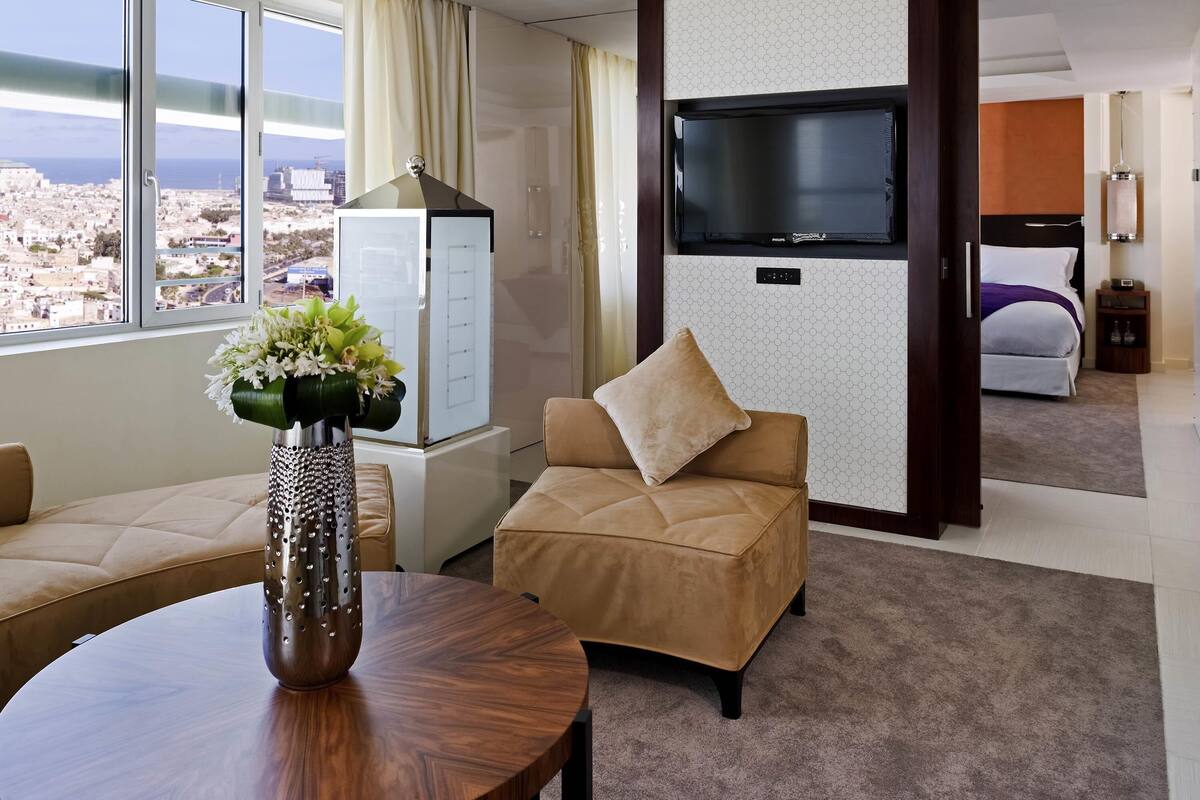 Habitación de hotel con sofá, una mesa redonda, televisor montado en la pared, cama y vista a la ciudad.