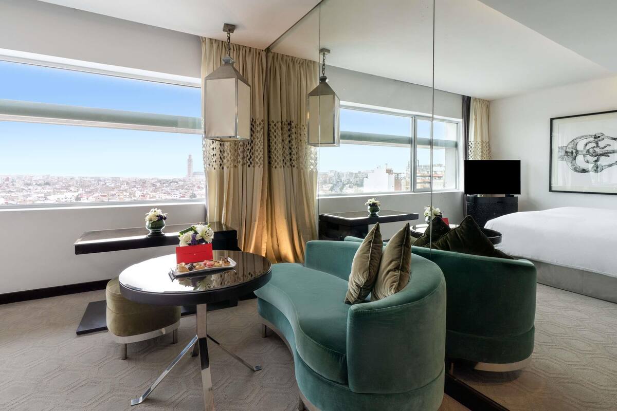 Una habitación de hotel moderna con una cama grande, dos sillones verdes, una mesita redonda con una bandeja de pasteles, un televisor de pantalla plana y vistas a la ciudad a través de amplias ventanas.