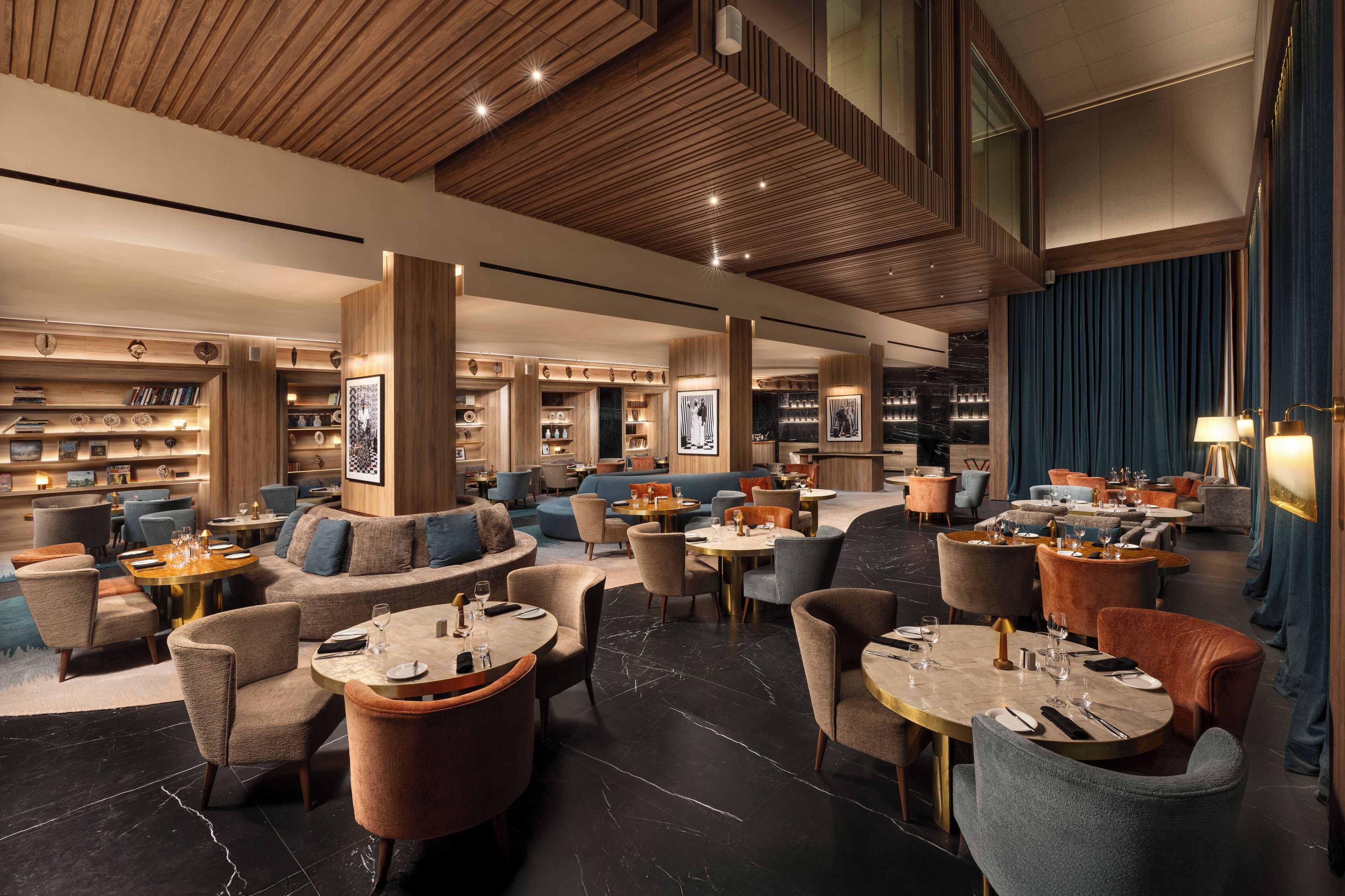 A spacious lounge area with multiple seating options including sofas and round tables, wooden paneling on the ceiling, and a large window with curtains.