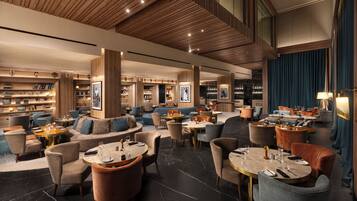 A spacious lounge area with multiple seating options including sofas and round tables, wooden paneling on the ceiling, and a large window with curtains.