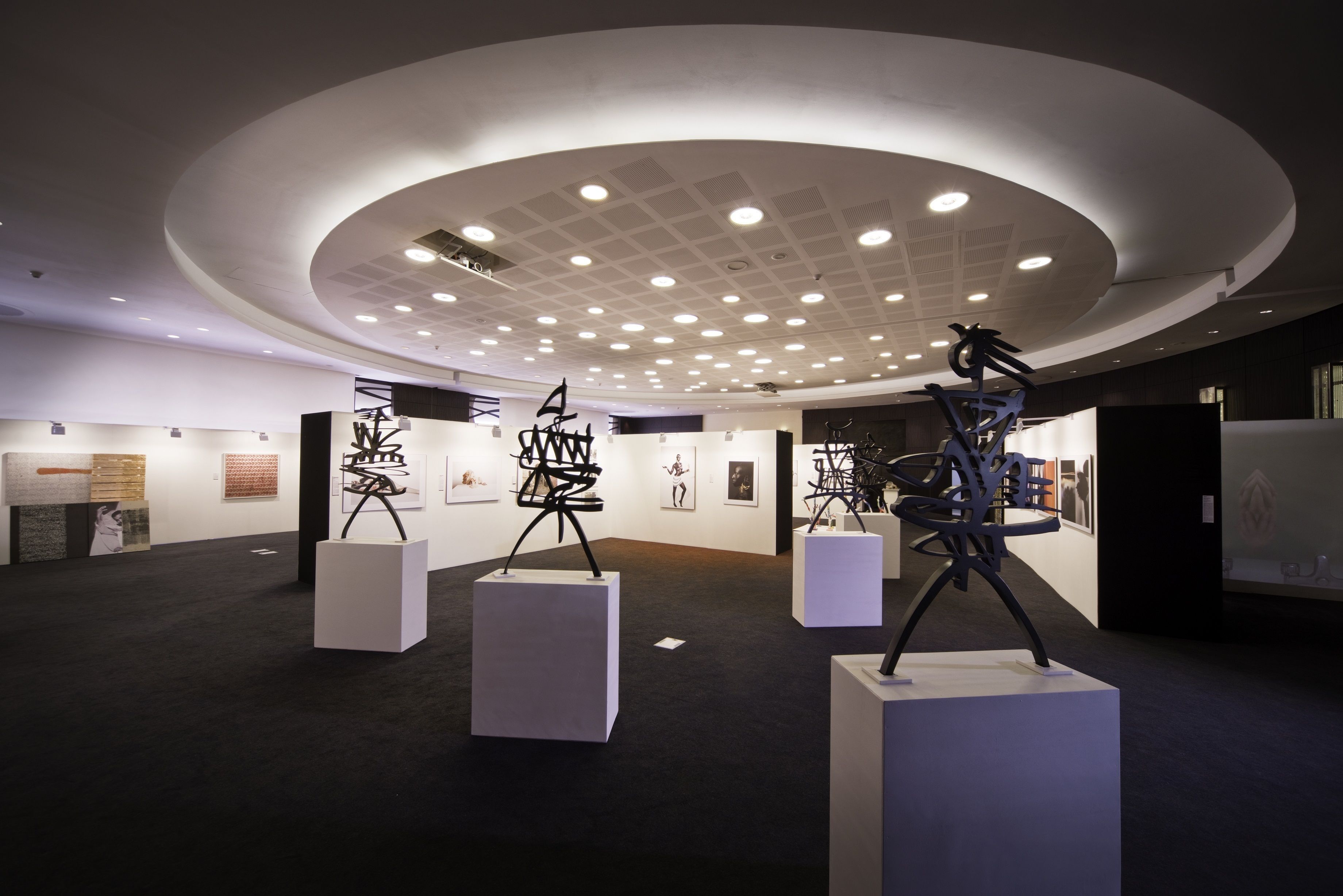 An art gallery with modern sculptures on display, white pedestals, and a ceiling with recessed lighting.