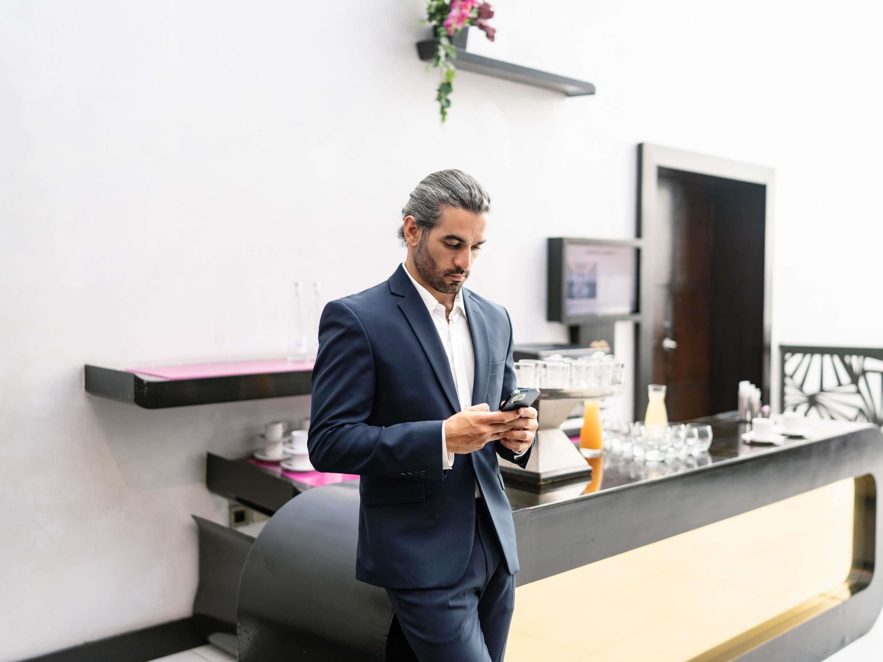 A man in a suit standing in a modern hotel lobby with a reception desk, a television, and a plant.
