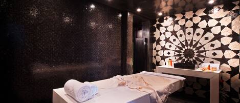 Une salle de spa équipée d’une table de massage, d’une serviette, d’une petite table avec des bouteilles et d’un mur décoré de motifs.