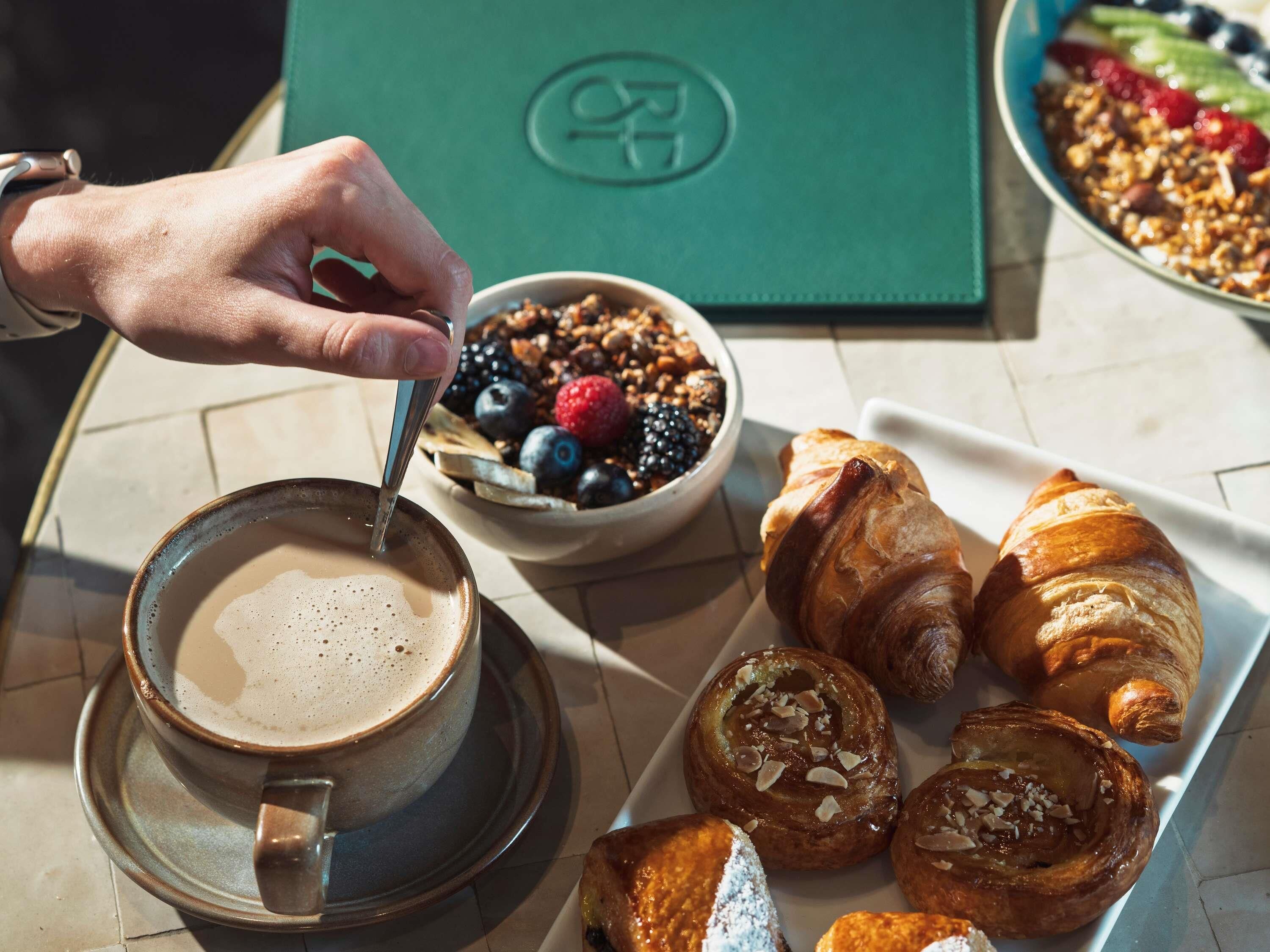 A breakfast setting with a cup of coffee, a bowl of granola with berries, and a plate of croissants.