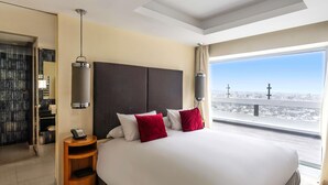 A hotel room with a large bed, a bedside table, a lamp, and a view of the city.