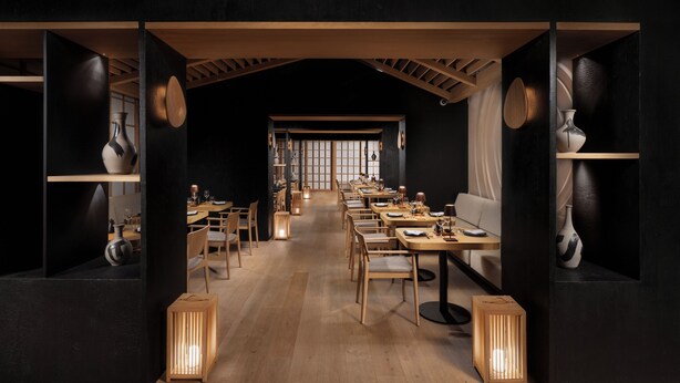Un restaurante tenuemente iluminado con mesas y sillas de madera, una barra larga y vistas a una habitación tradicional japonesa al fondo.