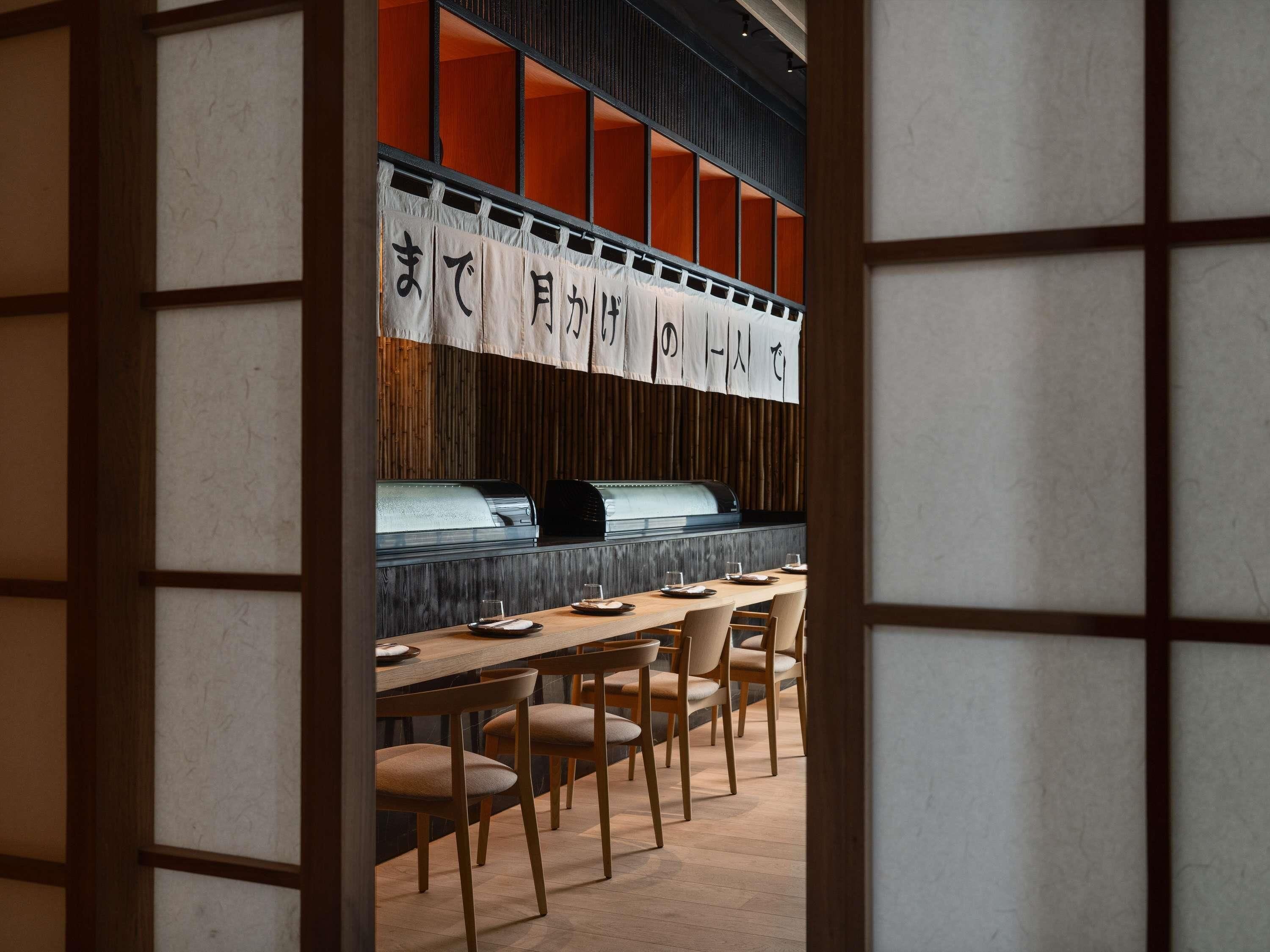 A restaurant interior with a long wooden table and chairs, a grill station, and a wall with Japanese characters.