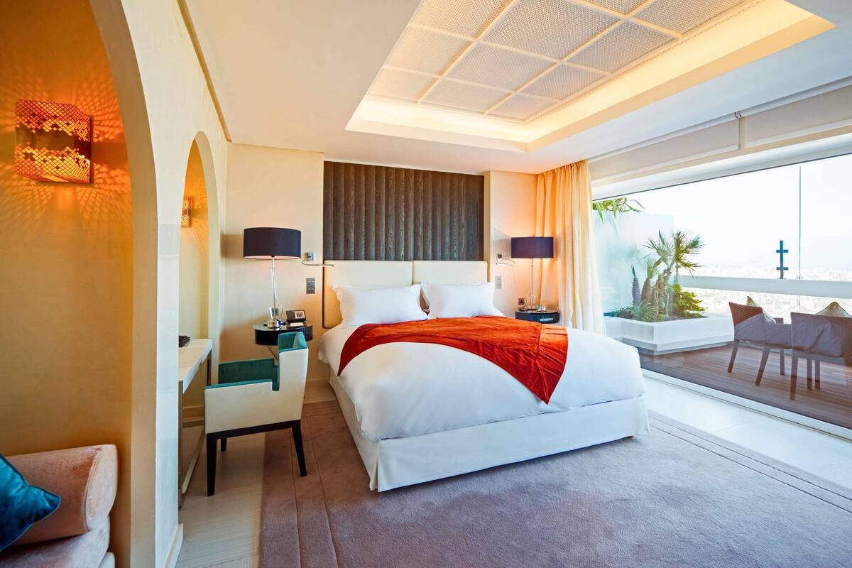 A modern hotel room with a large bed, a desk, and a balcony with seating.