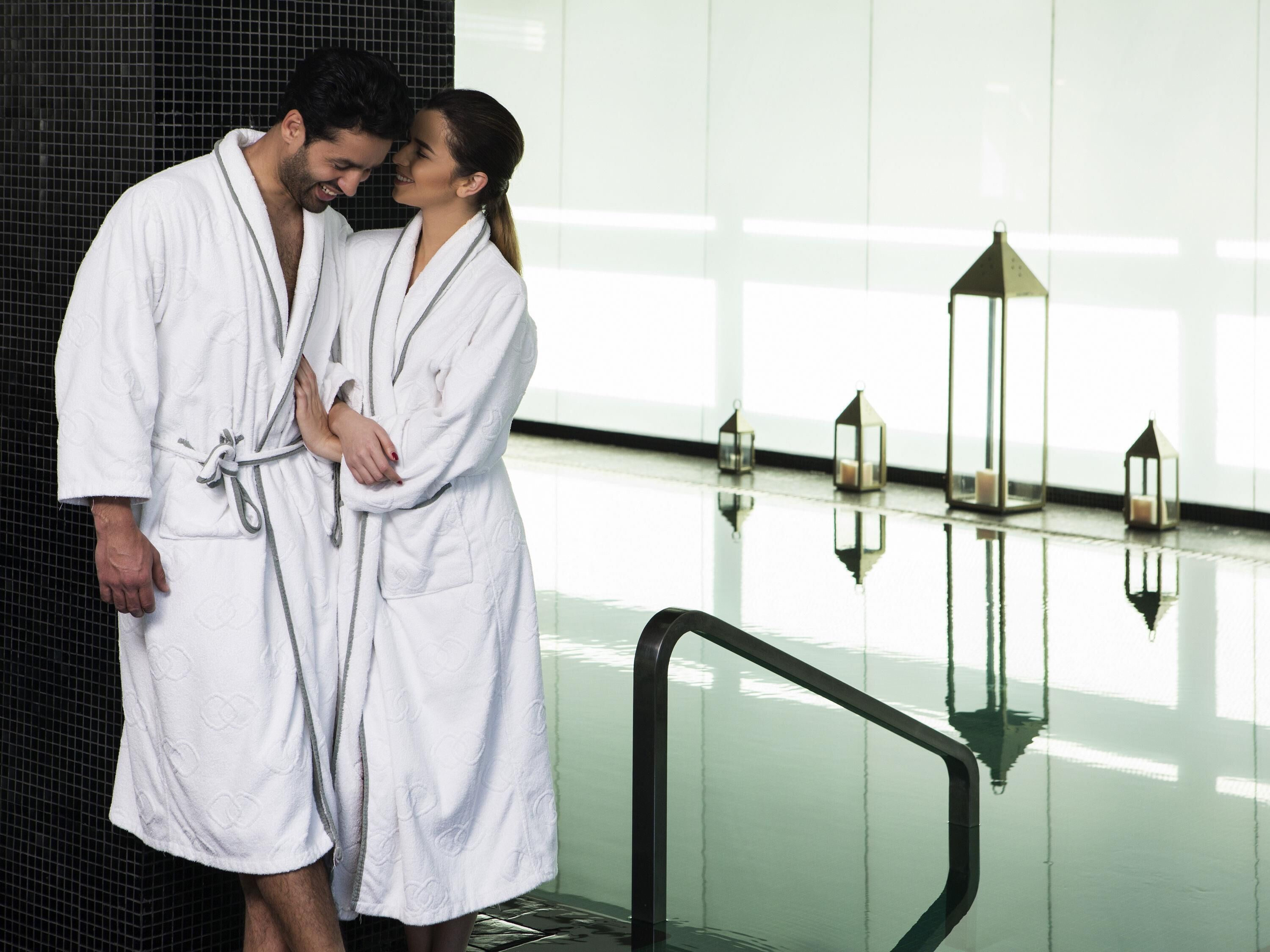 A couple in white bathrobes standing by a reflective pool with a glass railing and decorative lanterns.