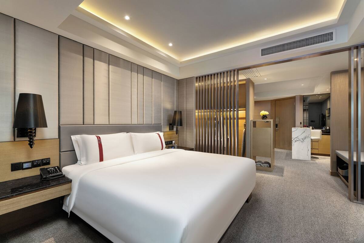 Une chambre d’hôtel moderne dotée d’un grand lit, de commandes situées près du lit, d’un bureau avec une lampe et d’une salle de bain visible à travers une cloison.