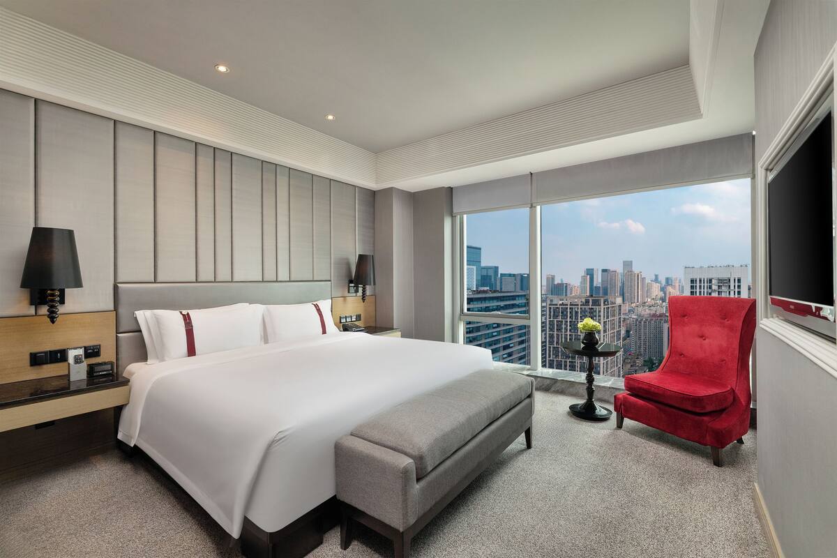 Une chambre d’hôtel moderne avec un grand lit, un fauteuil rouge, une vue sur la ville et un téléviseur à écran plat.