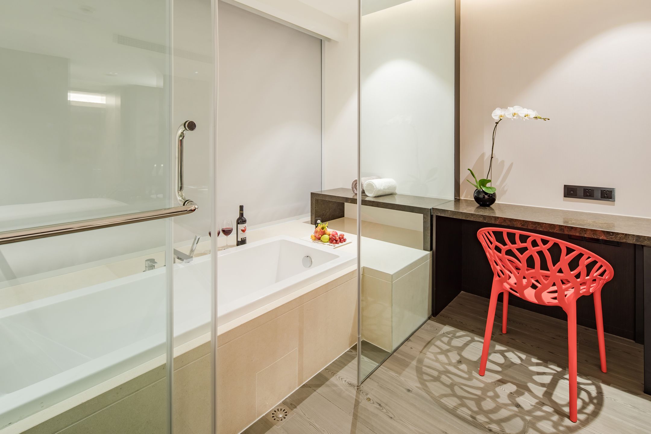 A modern bathroom with a glass shower enclosure, a bathtub, a vanity with a mirror, and a red chair.