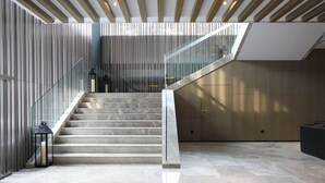 A modern interior with a staircase leading to a mezzanine, featuring a glass railing, wooden panel walls, and a polished stone floor.