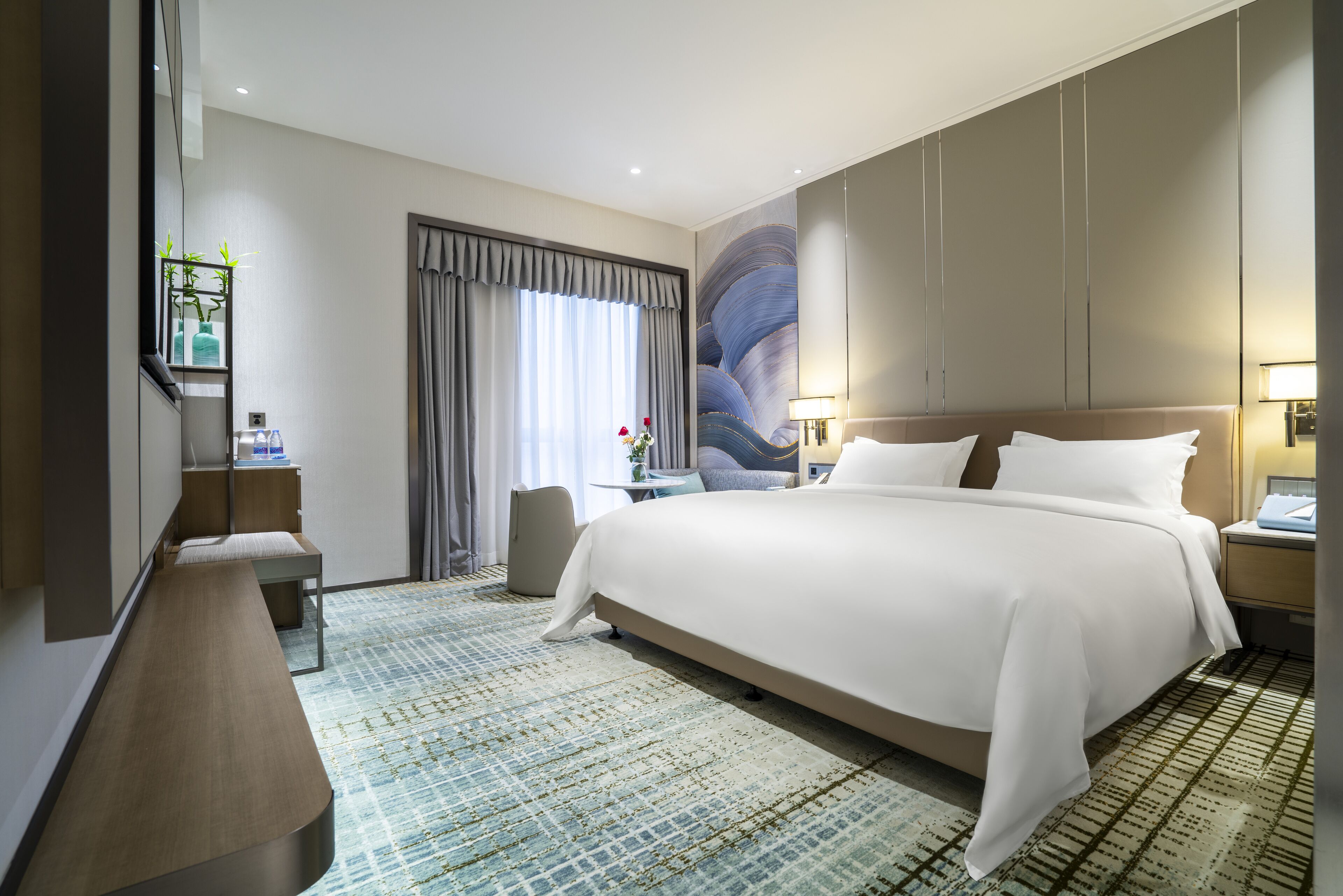 A modern hotel room with a large bed, a desk, a chair, and a large window with curtains.