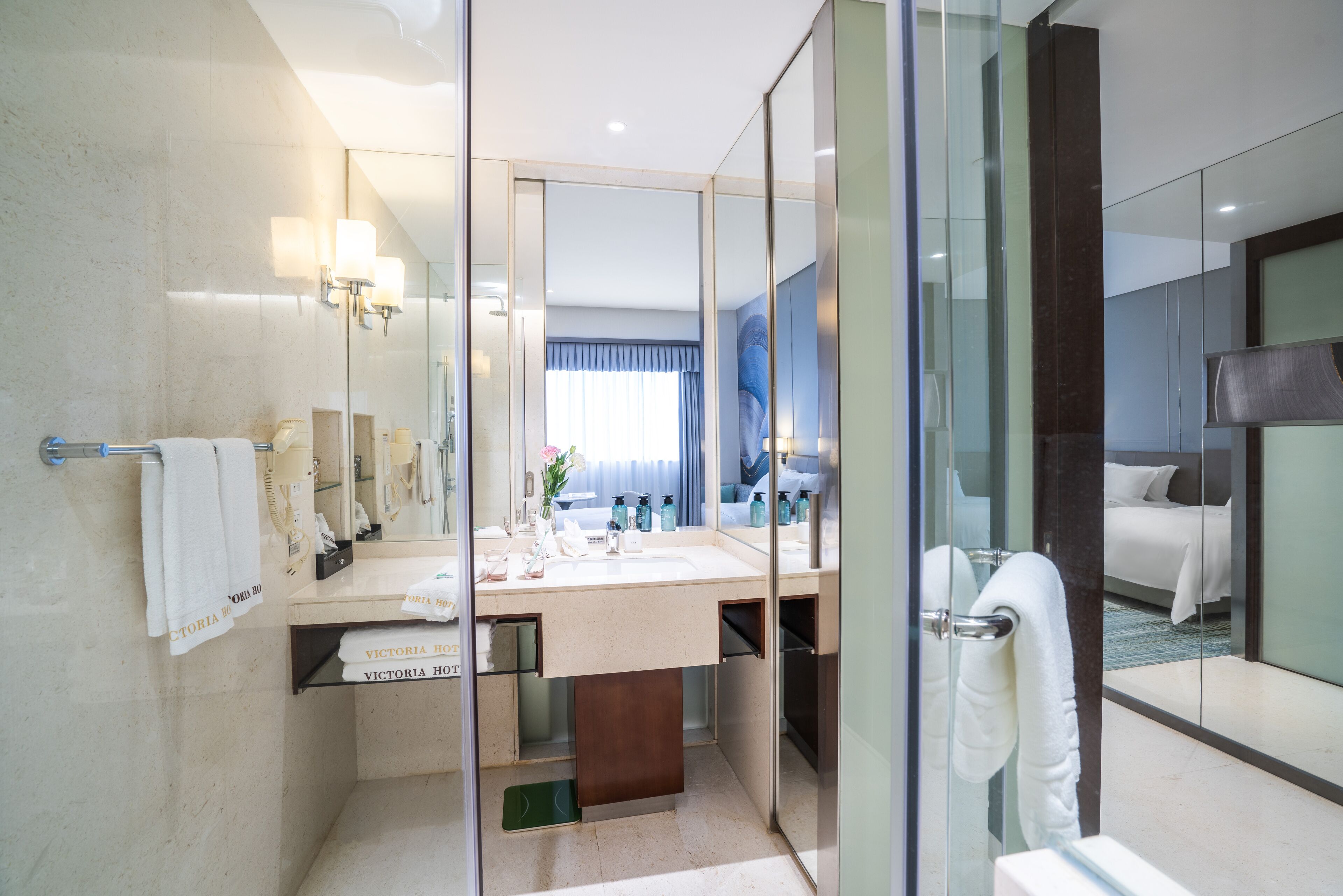 A modern hotel bathroom with a glass partition, a large mirror, a sink with toiletries, and a visible bed in the adjacent room.