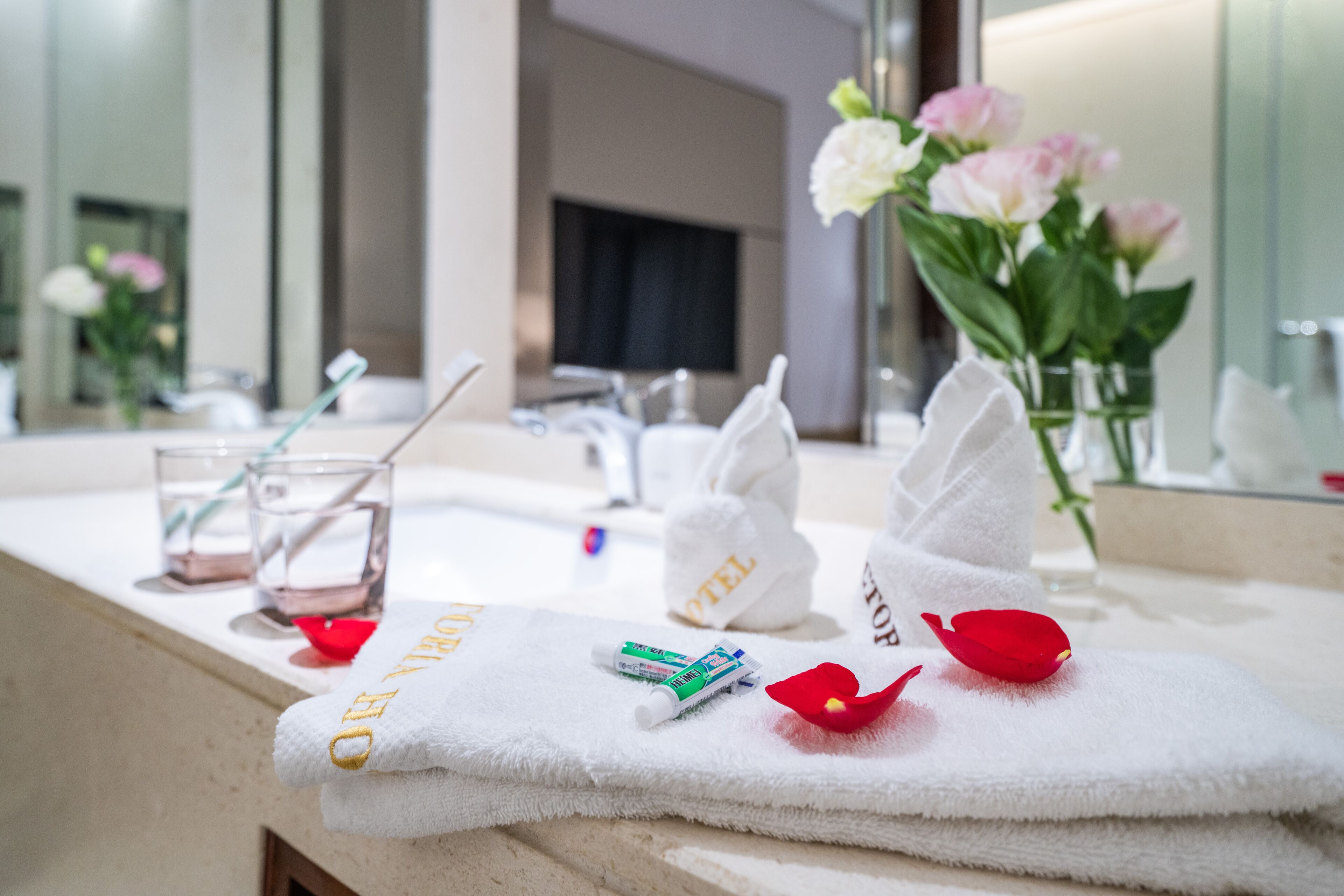Un baño de hotel con espejo, lavabo, toalla, vaso de agua, cepillo de dientes y un florero con flores.