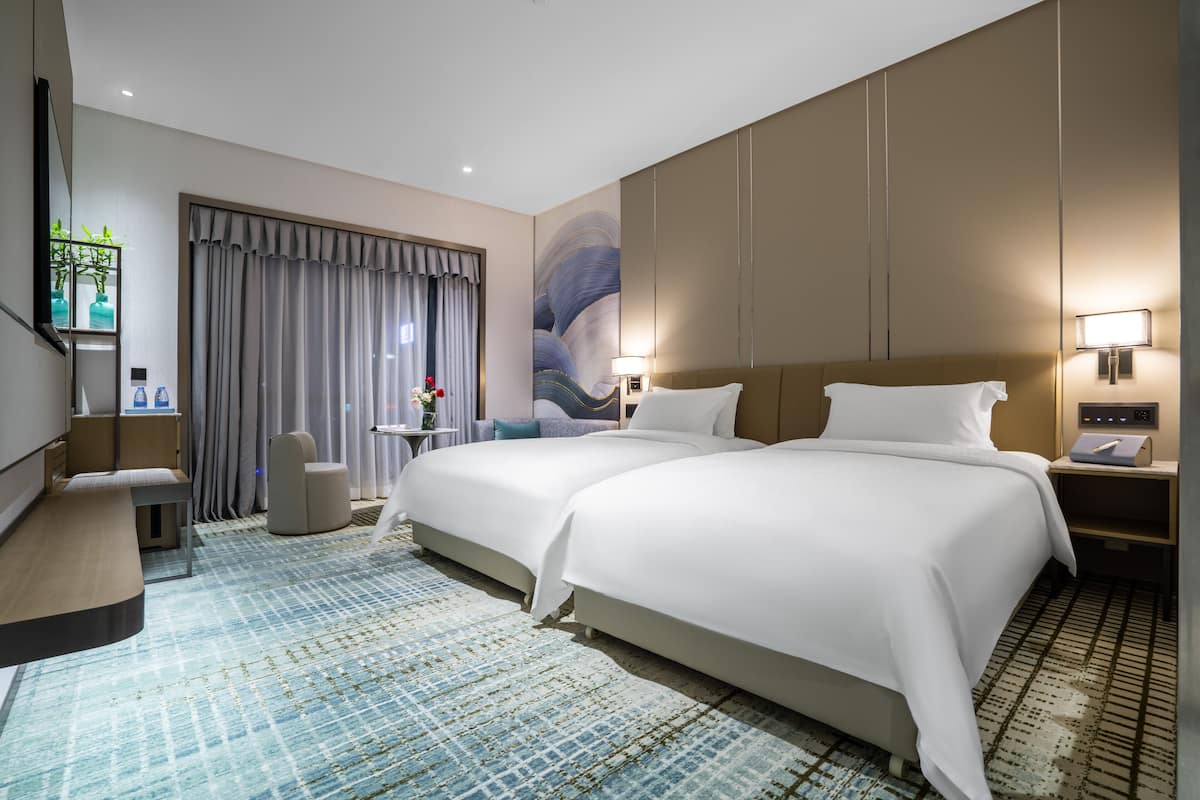 Habitación de hotel con dos camas, un escritorio, una silla, una mesita con un florero y un ventanal con cortinas.