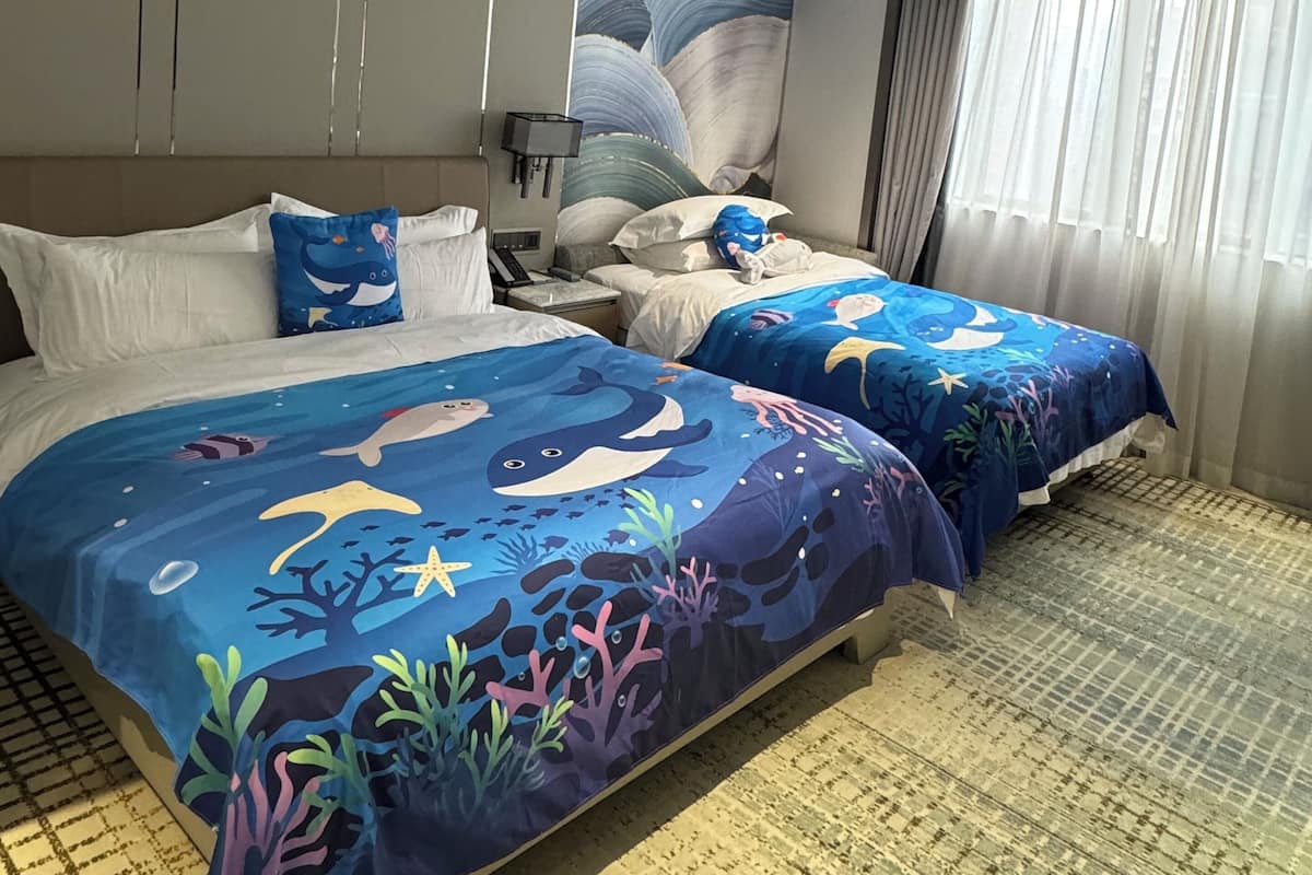 Habitación de hotel con dos camas, cada una con cubrecama de motivos marinos azules, un cuadro grande en la pared y vistas al exterior a través de una ventana con cortinas.