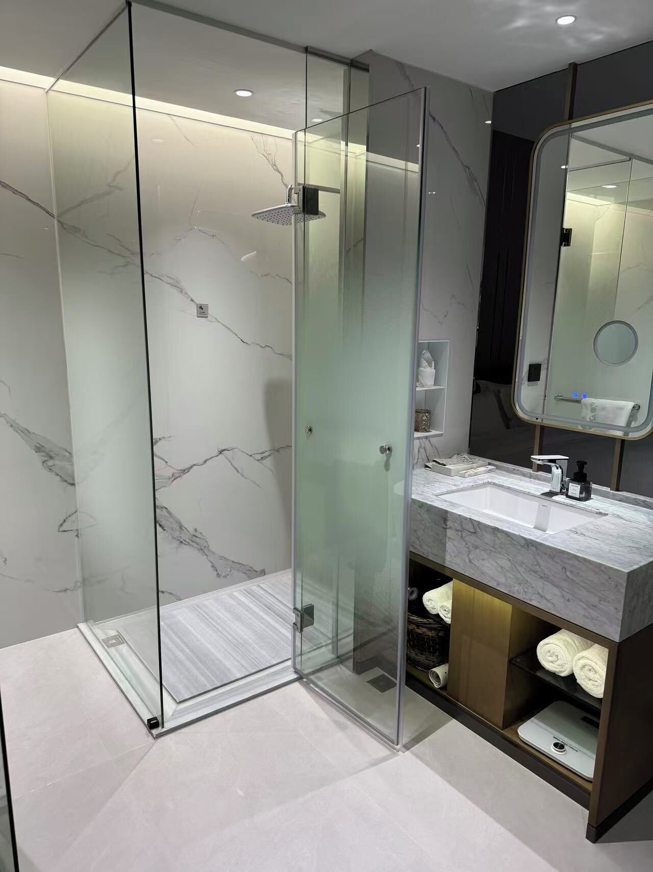 A modern bathroom with a glass-enclosed shower, marble-tiled walls, a vanity with a sink, and a mirror.