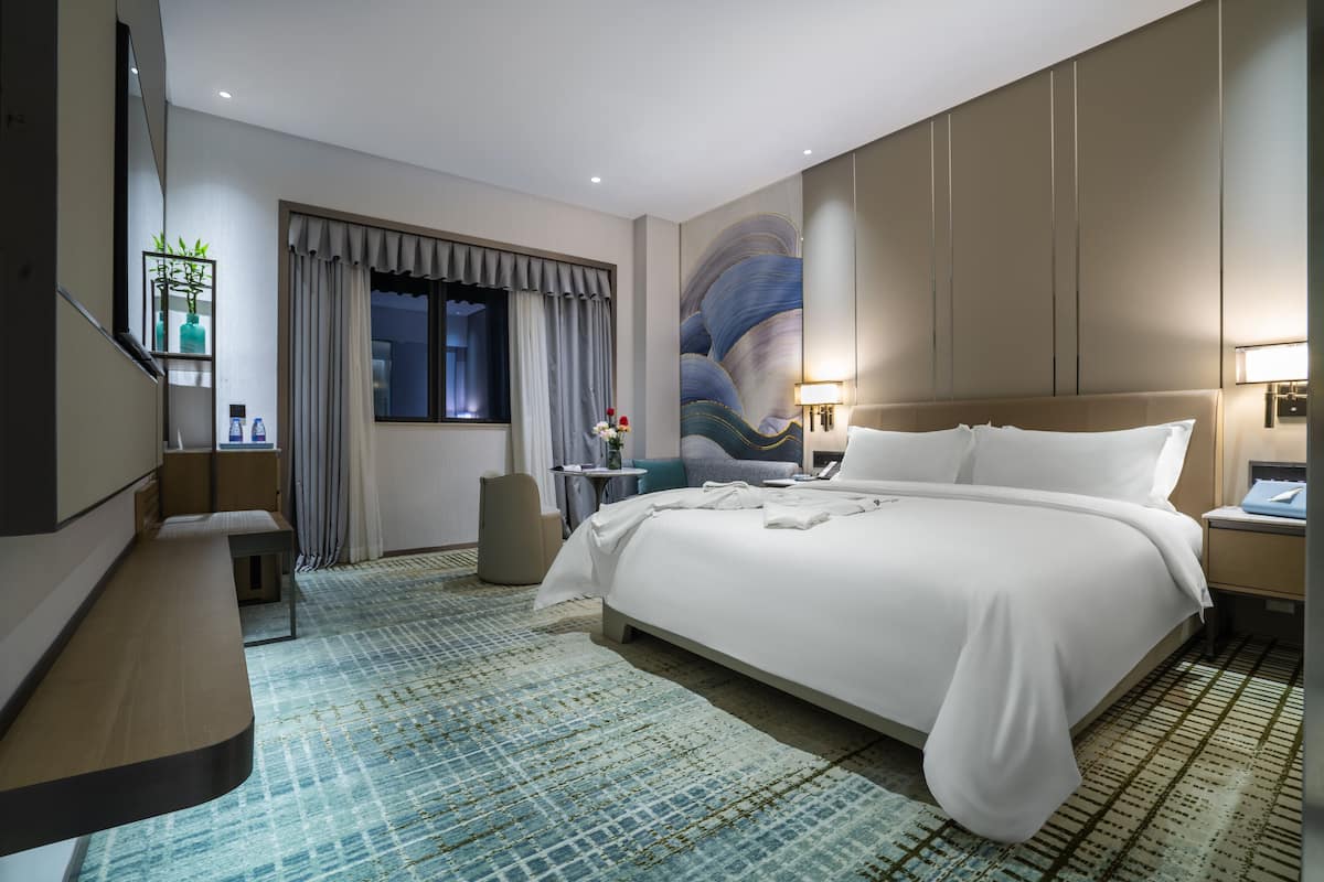 Una habitación de hotel moderna con una cama grande, una mesita de noche, un escritorio pequeño, una silla, un ventanal con cortinas, un mural en la pared y un área de baño con un espejo y estanterías.