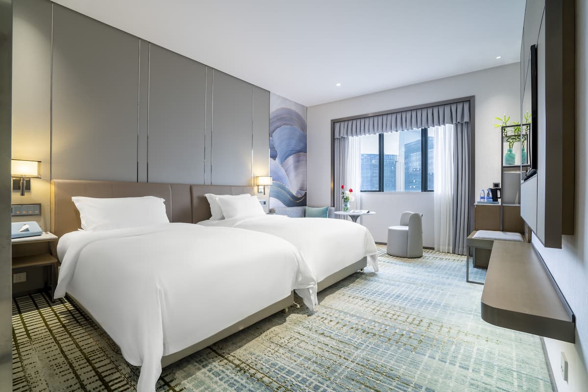 Habitación de hotel moderna con dos camas, un ventanal con cortinas, un escritorio y un televisor montado en la pared.