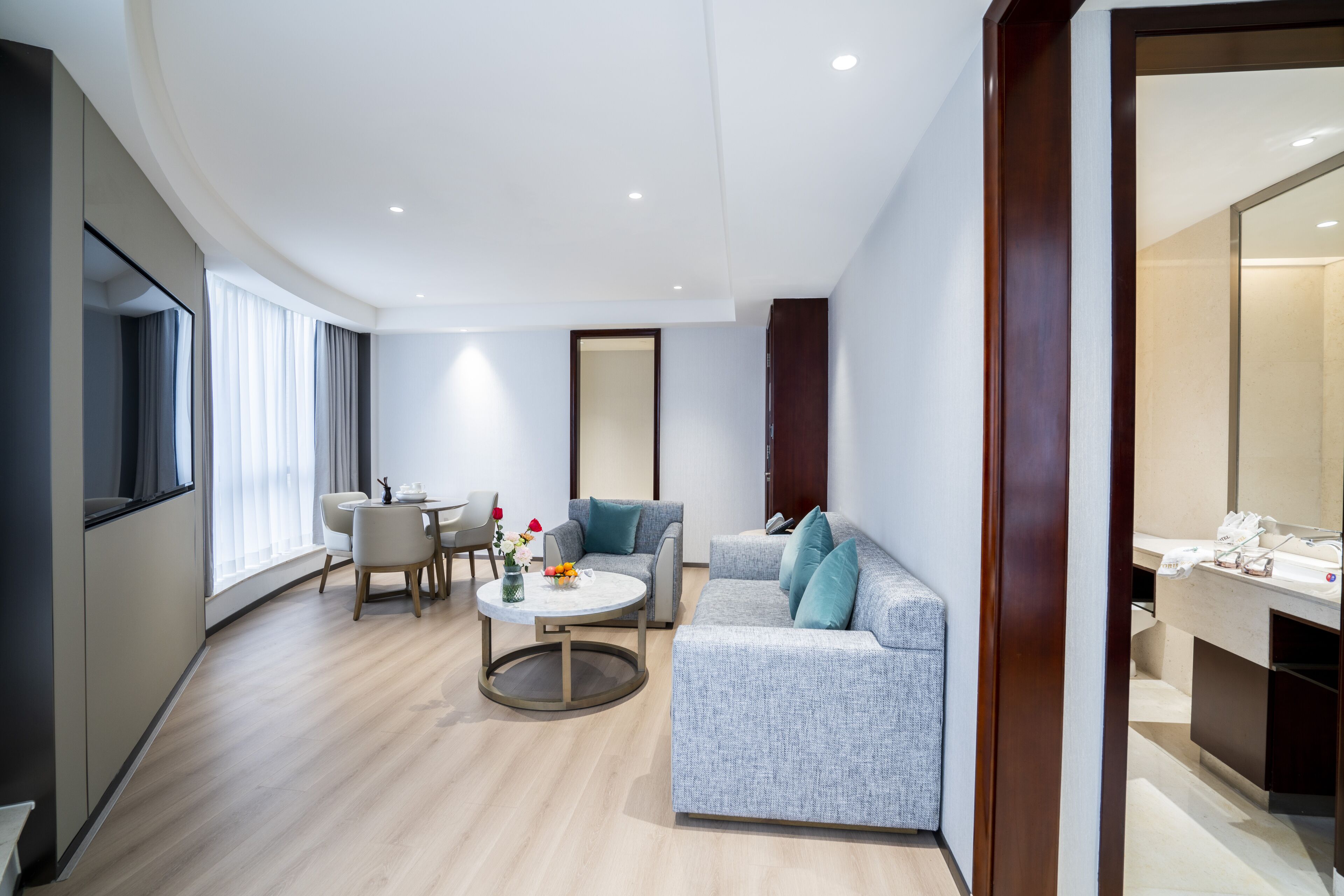 A modern hotel room with a grey sofa, a round coffee table, and a dining area with a table and chairs.
