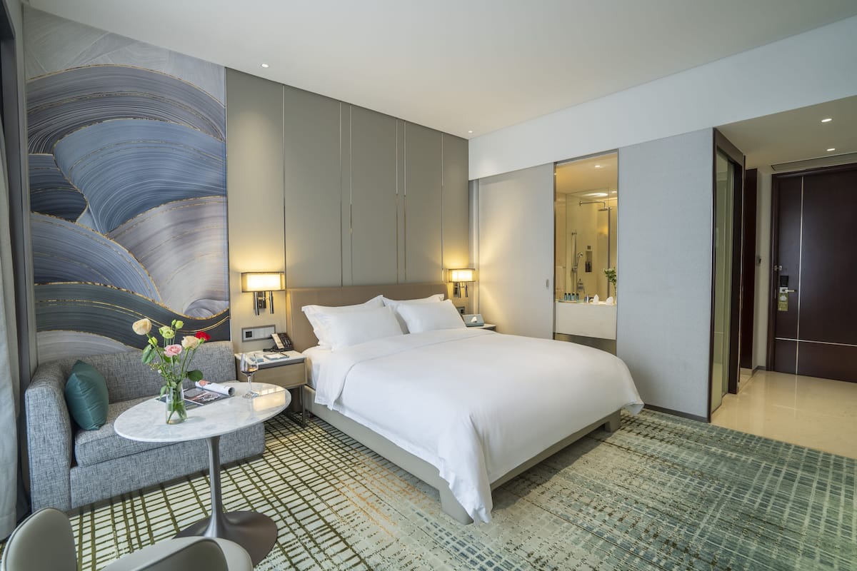 Una habitación de hotel moderna con una cama grande, un sofá, una pequeña mesa con un florero y un cuadro abstracto de gran tamaño en la pared.