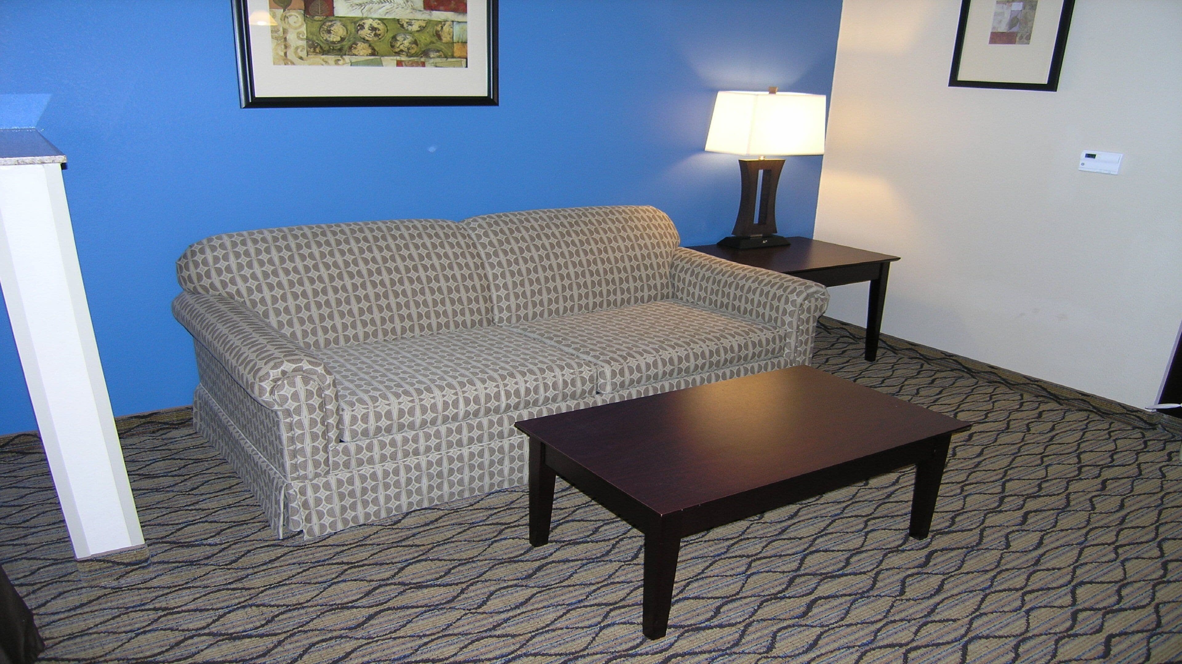 Une pièce avec un canapé à motifs, une petite table, une lampe et un sol recouvert d’une moquette.