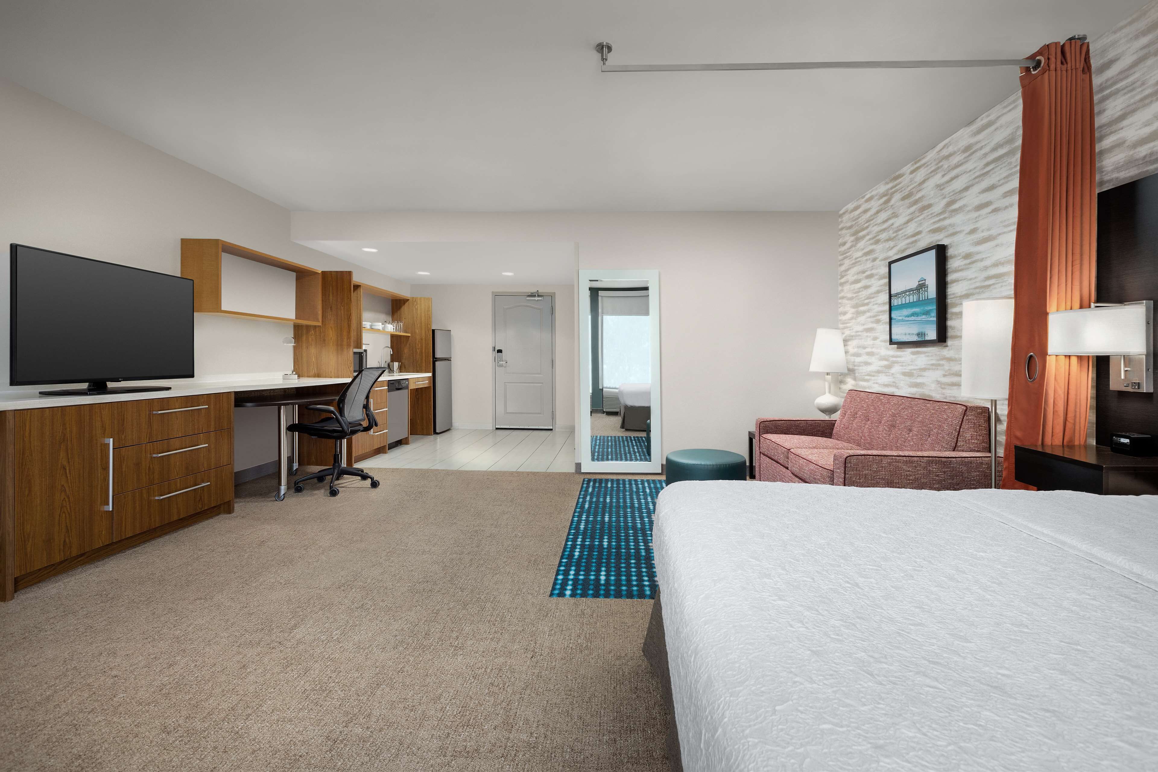 A modern hotel room with a large bed, a desk with a flat-screen TV, a kitchenette, a sofa, and a bathroom visible through an open door.