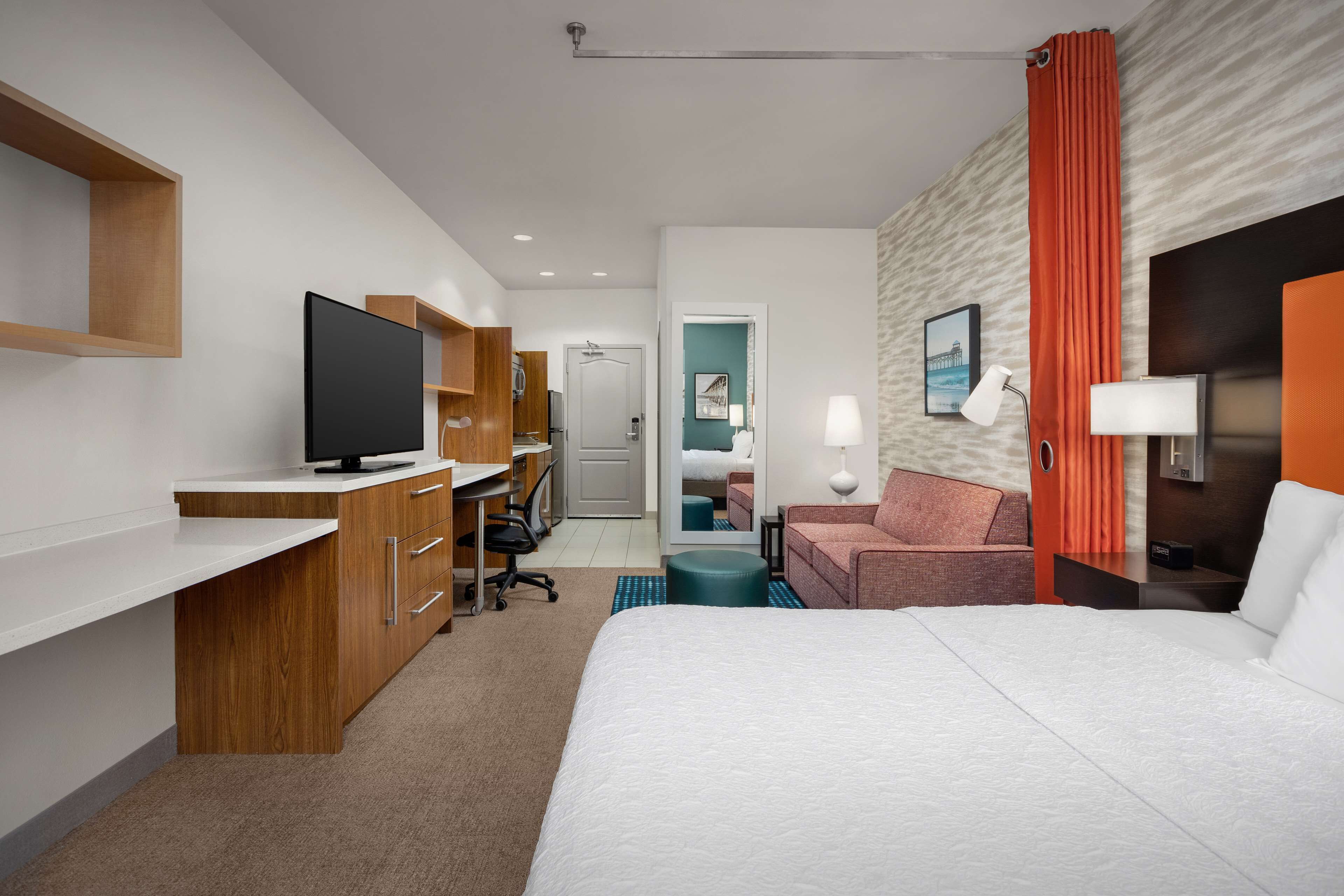 A hotel room with a bed, a desk with a TV, a chair, a sofa, and a bathroom visible through an open door.