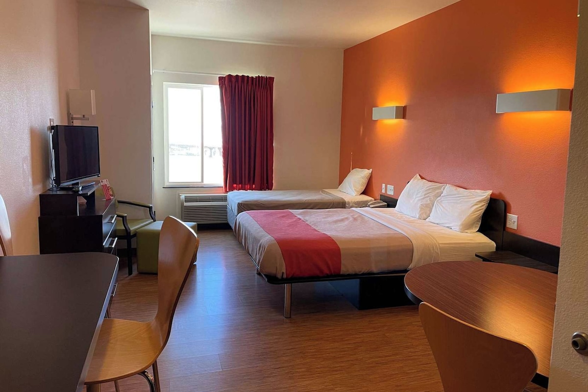 Habitación de hotel con dos camas, un escritorio, una silla, un televisor y una ventana con cortinas.