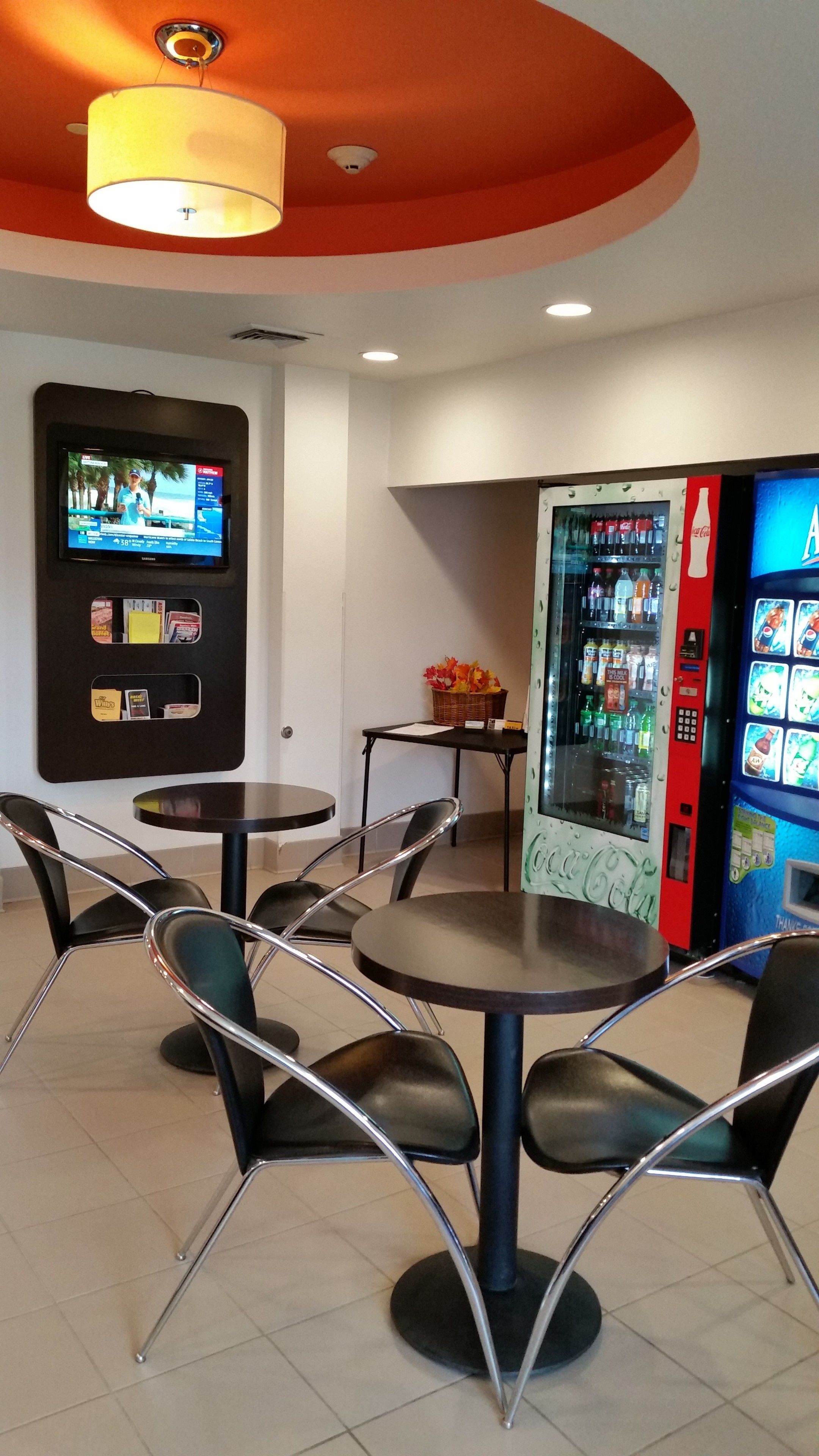 En restaurang med rundformade bord och stolar, en tv som hänger på väggen och en automattank för drycker.