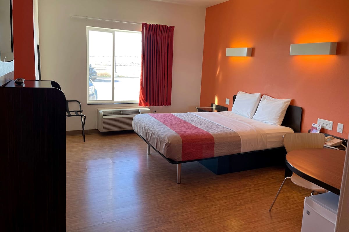 Una habitación de hotel con cama, escritorio, silla, ventana con cortinas e iluminación en la pared.