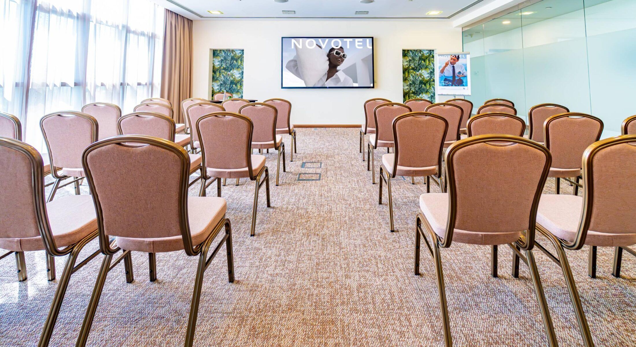 Uma sala de conferências com fileiras de cadeiras, piso coberto por carpete e uma tela grande exibindo 'NOVOTEL' na parede.