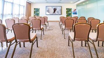 Uma sala de conferências com fileiras de cadeiras, piso coberto por carpete e uma tela grande exibindo 'NOVOTEL' na parede.