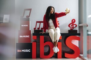 Una persona sentada en una plataforma con letras rojas grandes que dicen 'ibis MUSIC' y notas musicales al fondo.