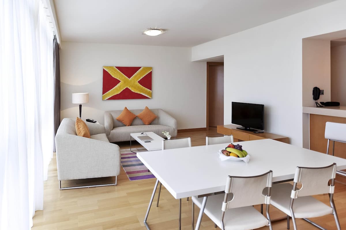 A modern living room with a sofa, a TV, a dining table with chairs, and a fruit bowl.