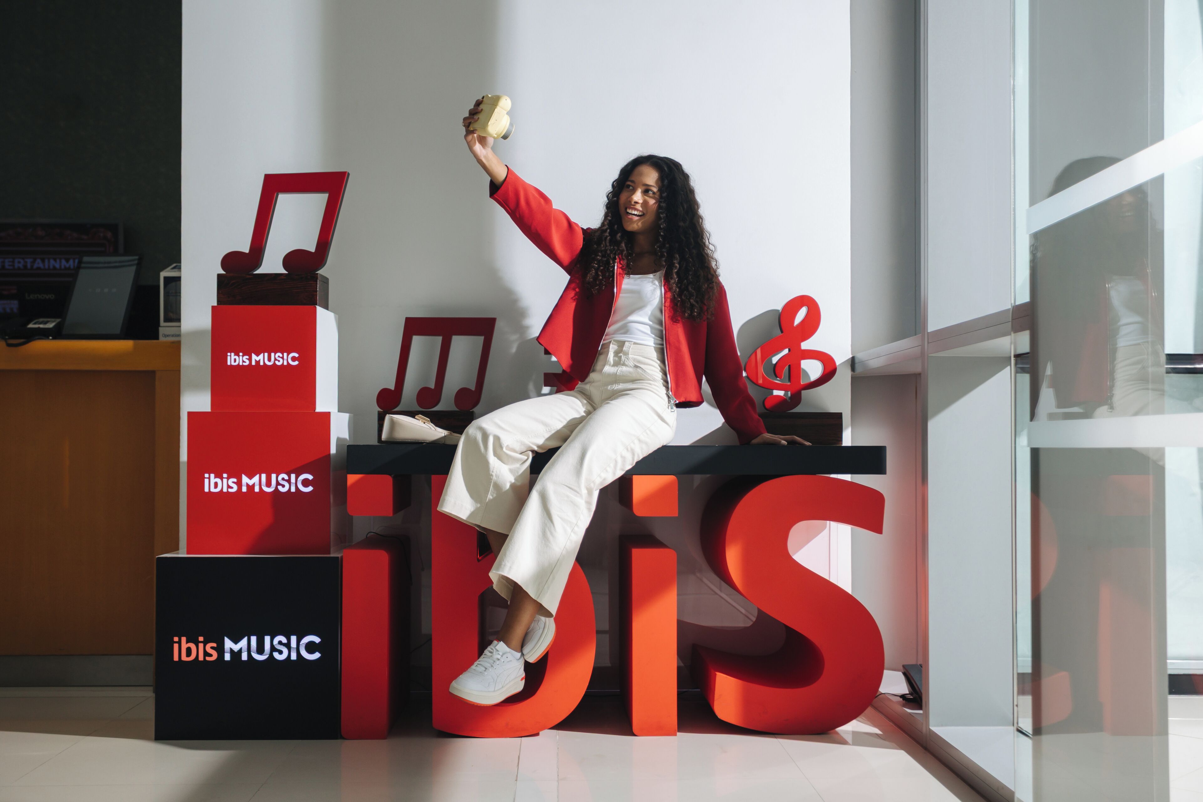 Una persona sentada sobre un gran logotipo rojo de 'ibis MUSIC', con una computadora portátil y una silla roja al fondo.