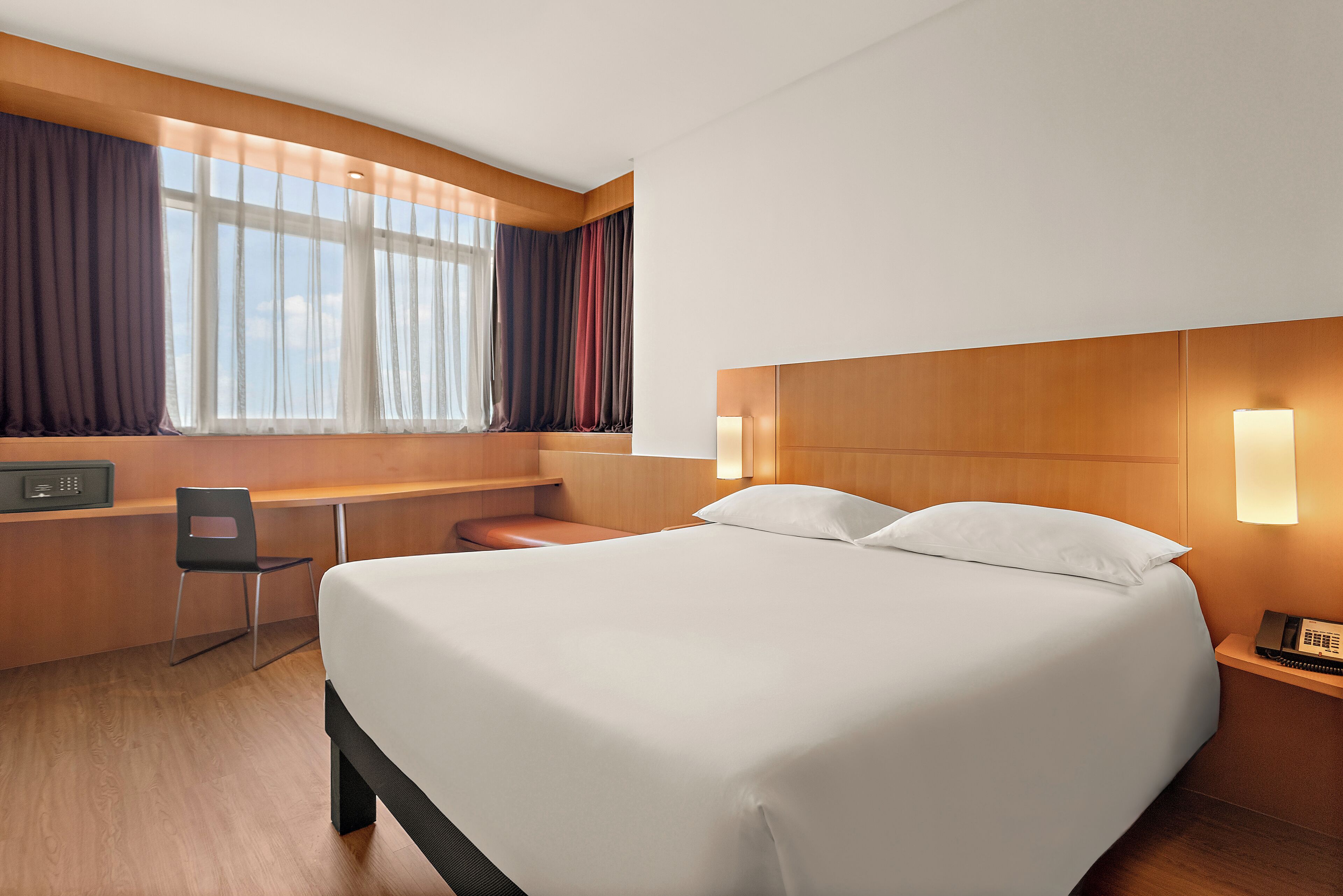 A hotel room with a large bed, a desk with a chair, a telephone, and a window with curtains.