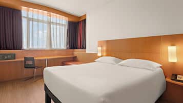 A hotel room with a large bed, a desk with a chair, a telephone, and a window with curtains.