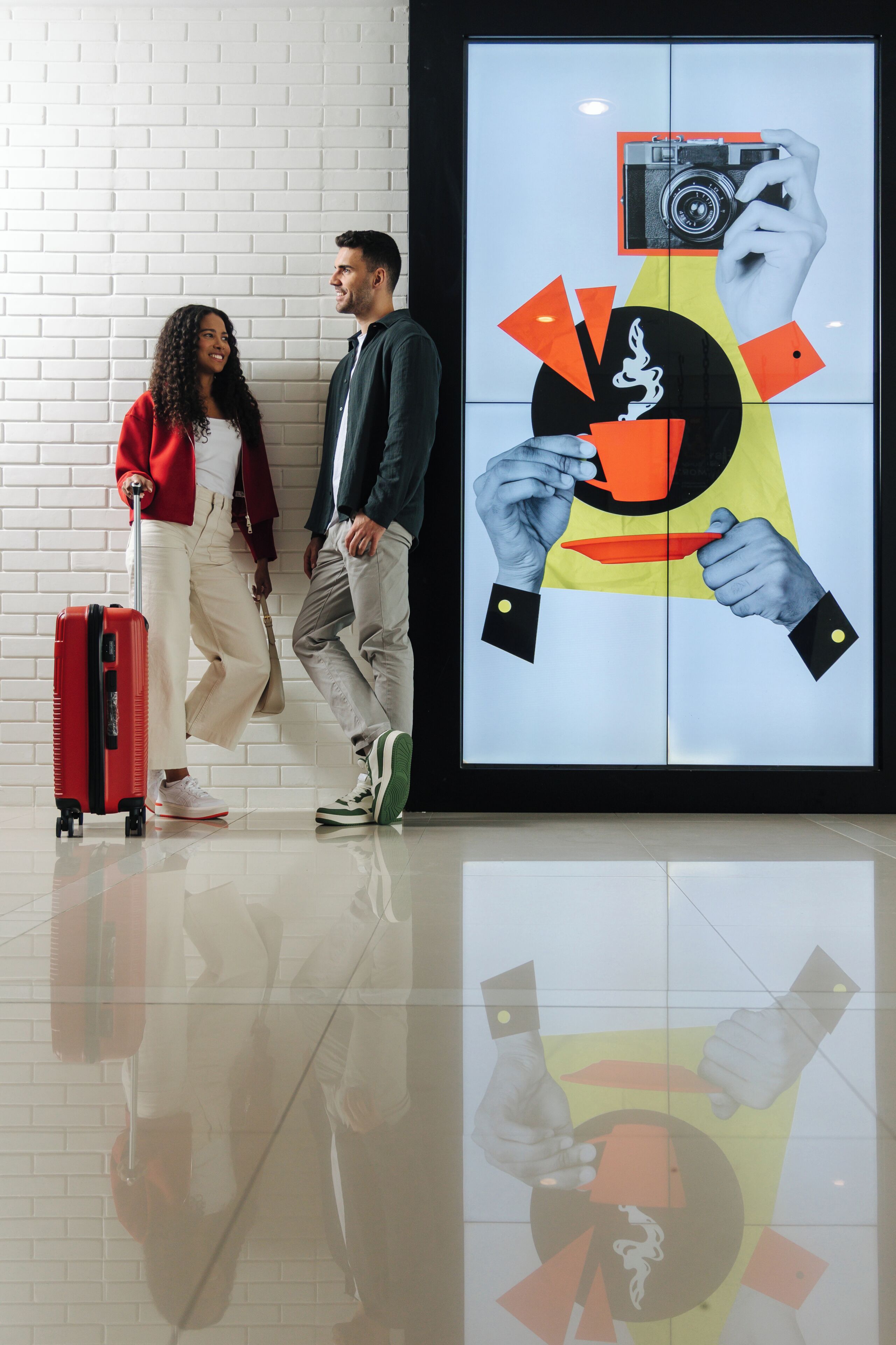 A couple with luggage, a red suitcase, and a black camera in a modern lobby with a tiled floor.